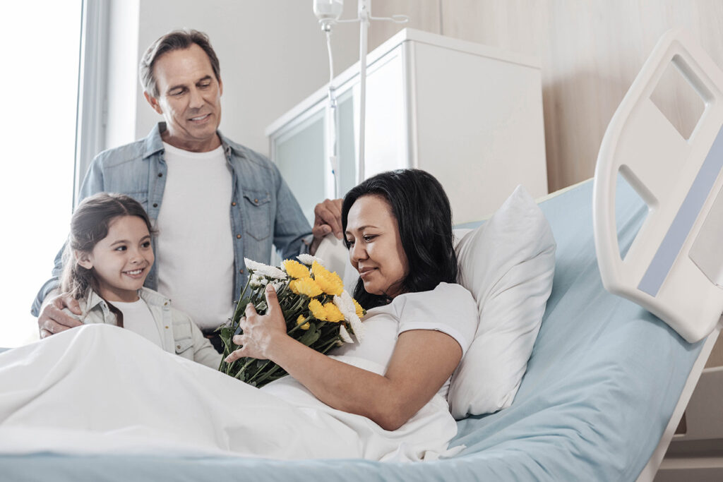 Une mère est à l'hôpital et son mari ainsi que sa fille viennent lui apporter des fleurs lors d'une visite.